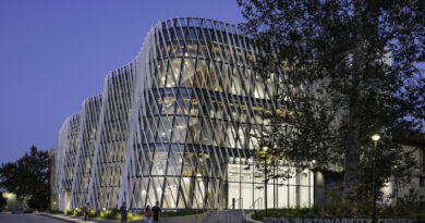 A photo of the entrance of the Resnick Sustainability Center at California Institute of Technology. The building is transparent with lights illuminating the inside. A sign reads the name of the building and is next to a large tree.