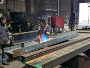 Two individuals in a warehouse working with steel fabrication tools.