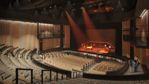 Rendering of a large auditorium with students performing for a class on the stage under warm orange spot lights.